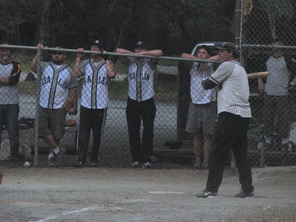 Baseball team in uniforms standing behind a chain-link fence, listening to a coach on the field.