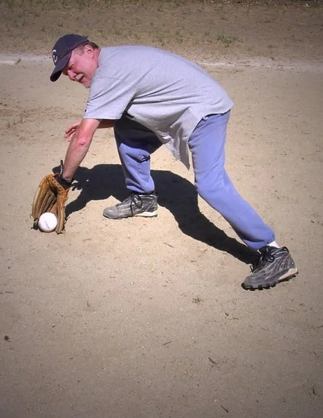 Man playing baseball, crouched down in the dirt in a field with a baseball glove and ball.