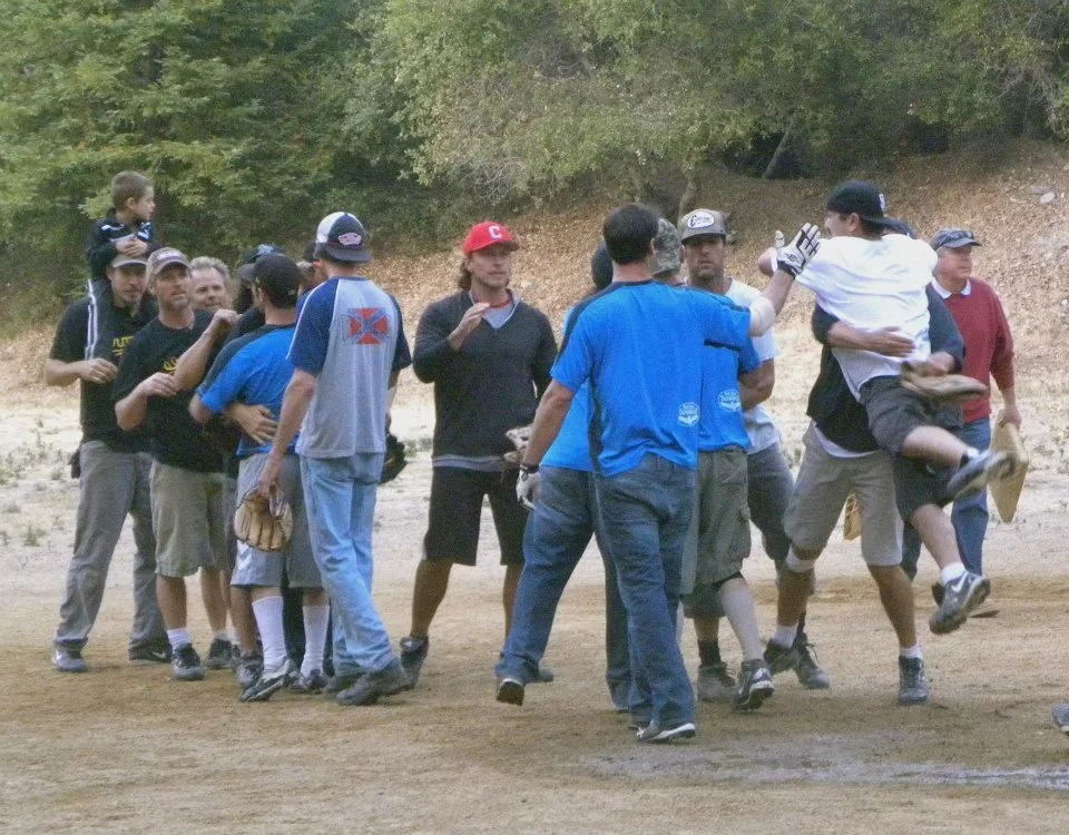 A group of young men and boys engaging in a baseball game, with some spectators in the background, on a dirt field with trees.