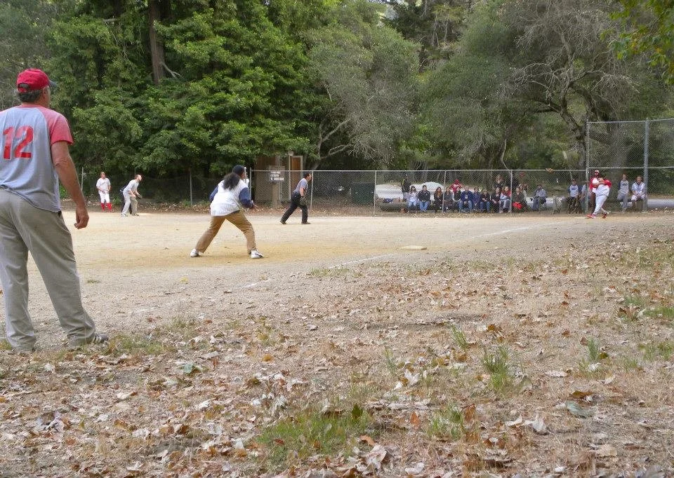 People playing a baseball game on a dirt field with spectators sitting on benches behind a chain-link fence, surrounded by trees.