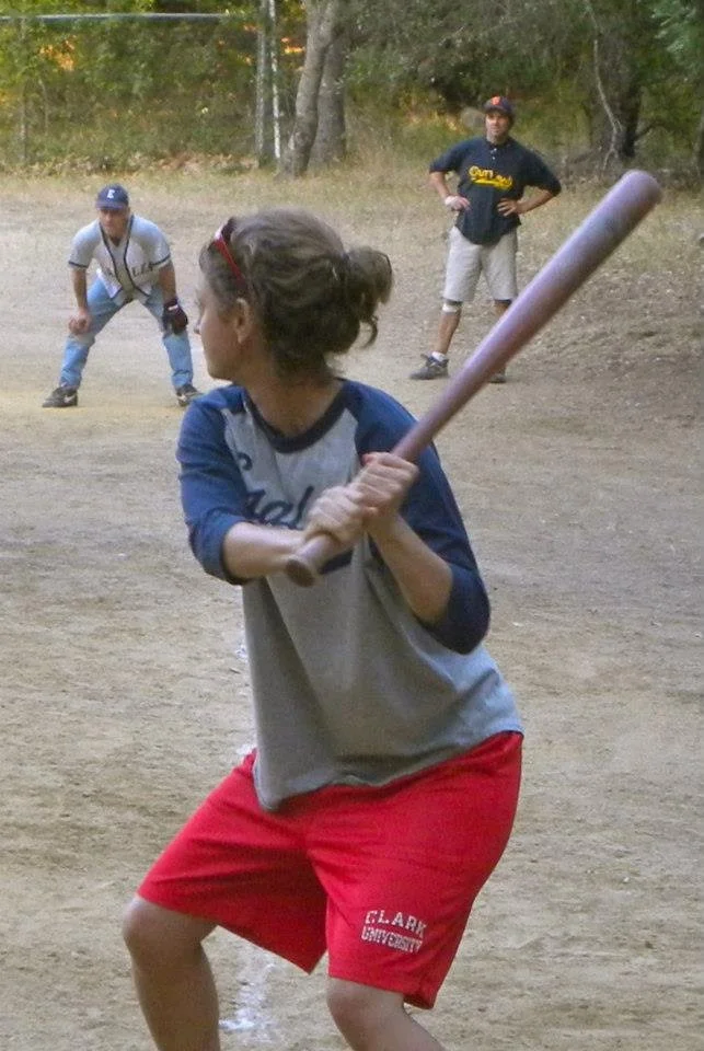 A young girl in a baseball uniform holding a bat while preparing to swing, on a baseball field with two women observing in the background.