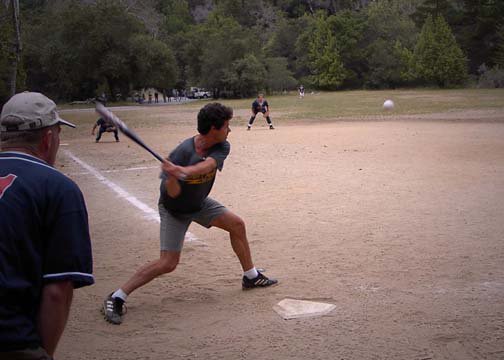 A man swinging a bat at a baseball on a dirt field, with players and spectators in the background.