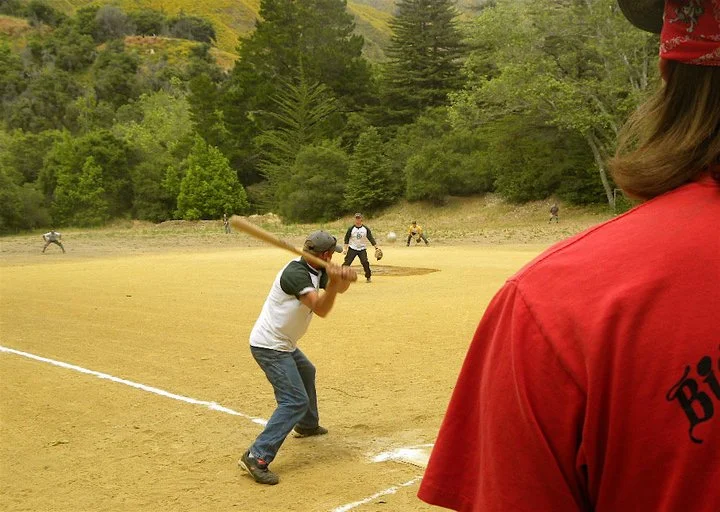 Kids playing baseball on a dirt field surrounded by trees, with some people in the background.