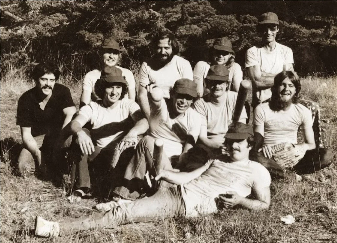 Group of ten people outdoors, smiling, some wearing hats, some holding a baseball bat, in a grassy area with trees in the background.