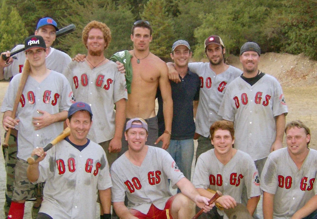 Group of people in baseball uniforms and casual clothing posing outdoors; some holding baseball bats; trees and dirt field in background.
