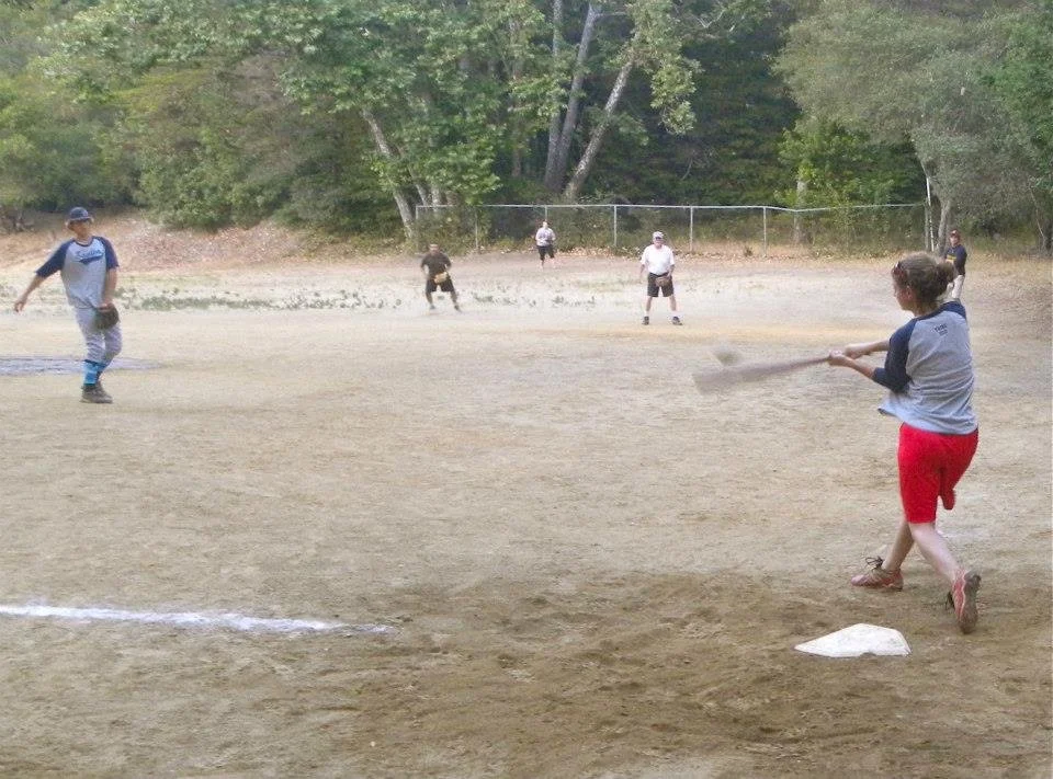 Children playing baseball on a dirt field with trees in the background.