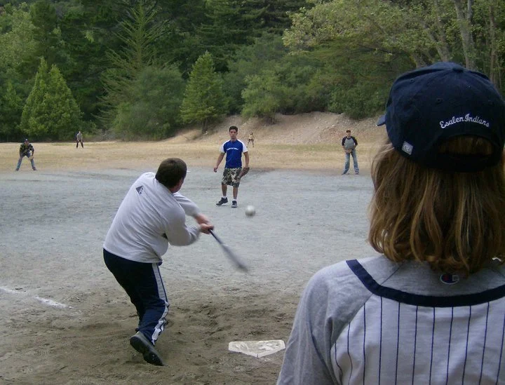 Children playing baseball outdoors on a dirt field during daytime, with trees and hills in the background.