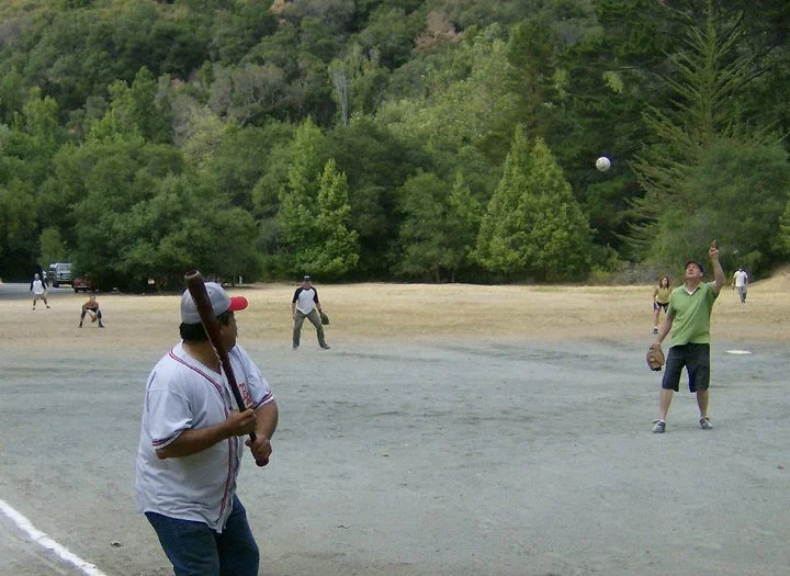People playing baseball on a field with a backdrop of green trees and hills.