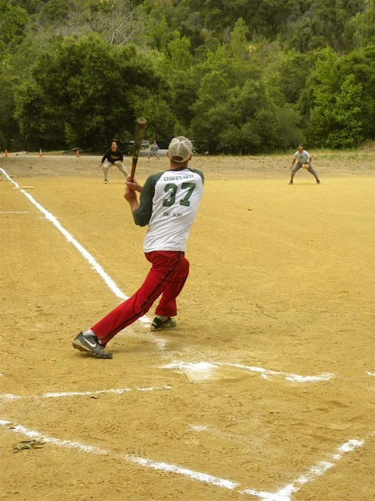 A person in a green shirt and red pants batting during a baseball game on a dirt field, with two players in the background ready to catch, surrounded by trees.