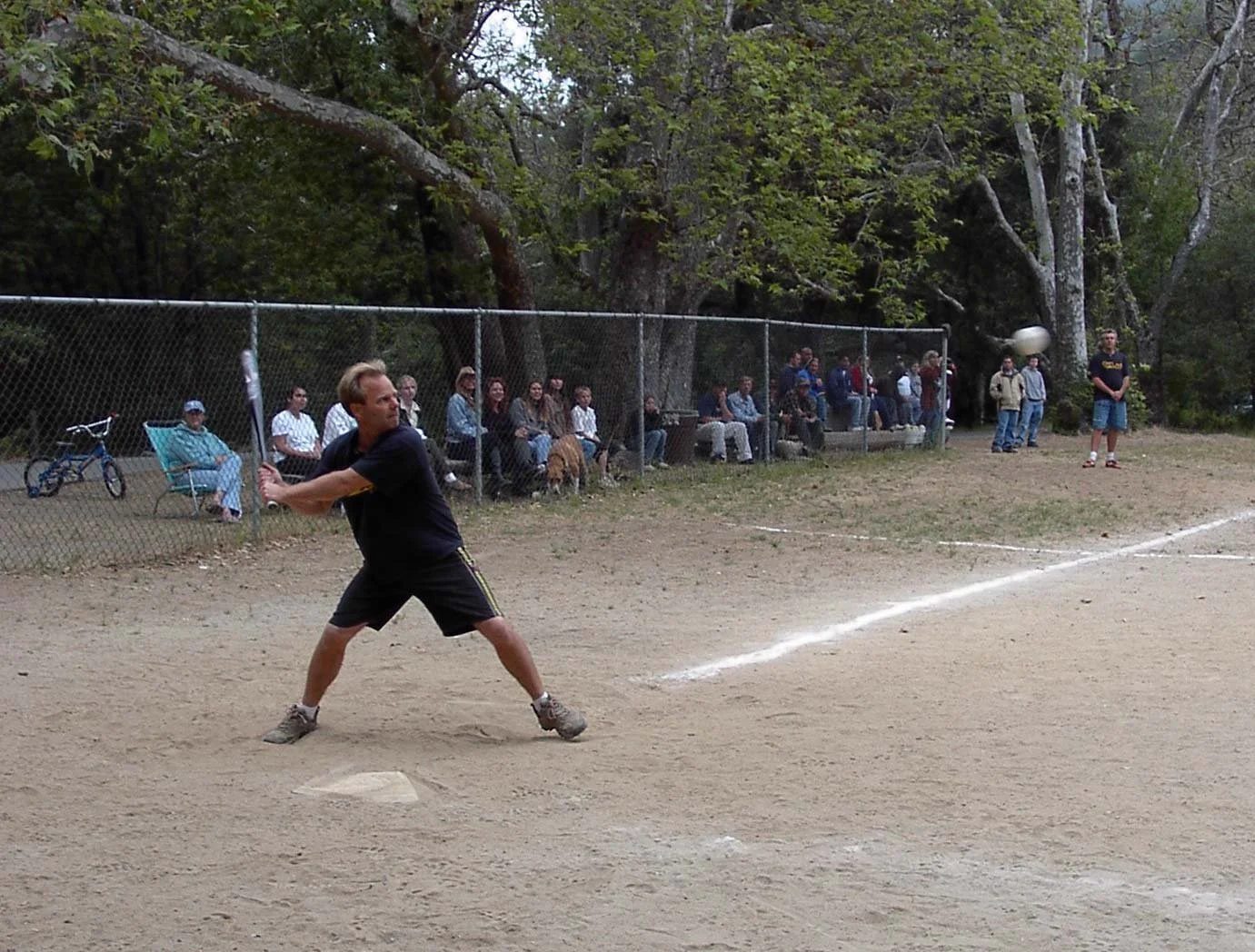 A man in black shorts and a black T-shirt playing baseball on a dirt field, preparing to hit a pitched ball, with spectators seated and standing behind a chain-link fence in a wooded park.