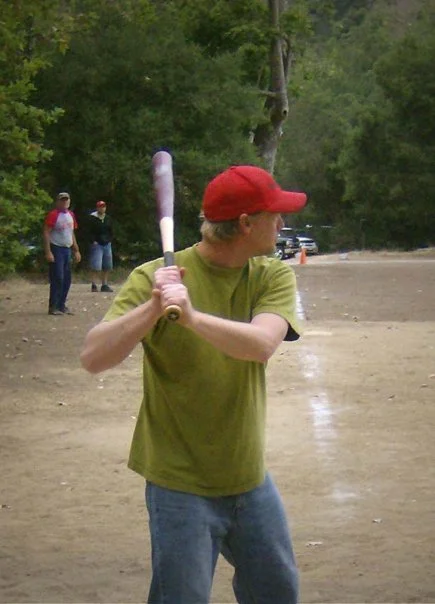 Teenager in a red cap and green t-shirt holding a baseball bat on a baseball field with two people in the background.