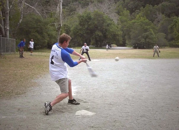 A young man batting at a baseball game on a gravel field with several other players and trees in the background.