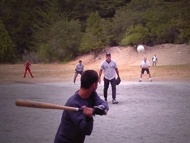 A group of men playing baseball on a field with trees and hills in the background. One man is swinging a bat; other men are positioned defensively.