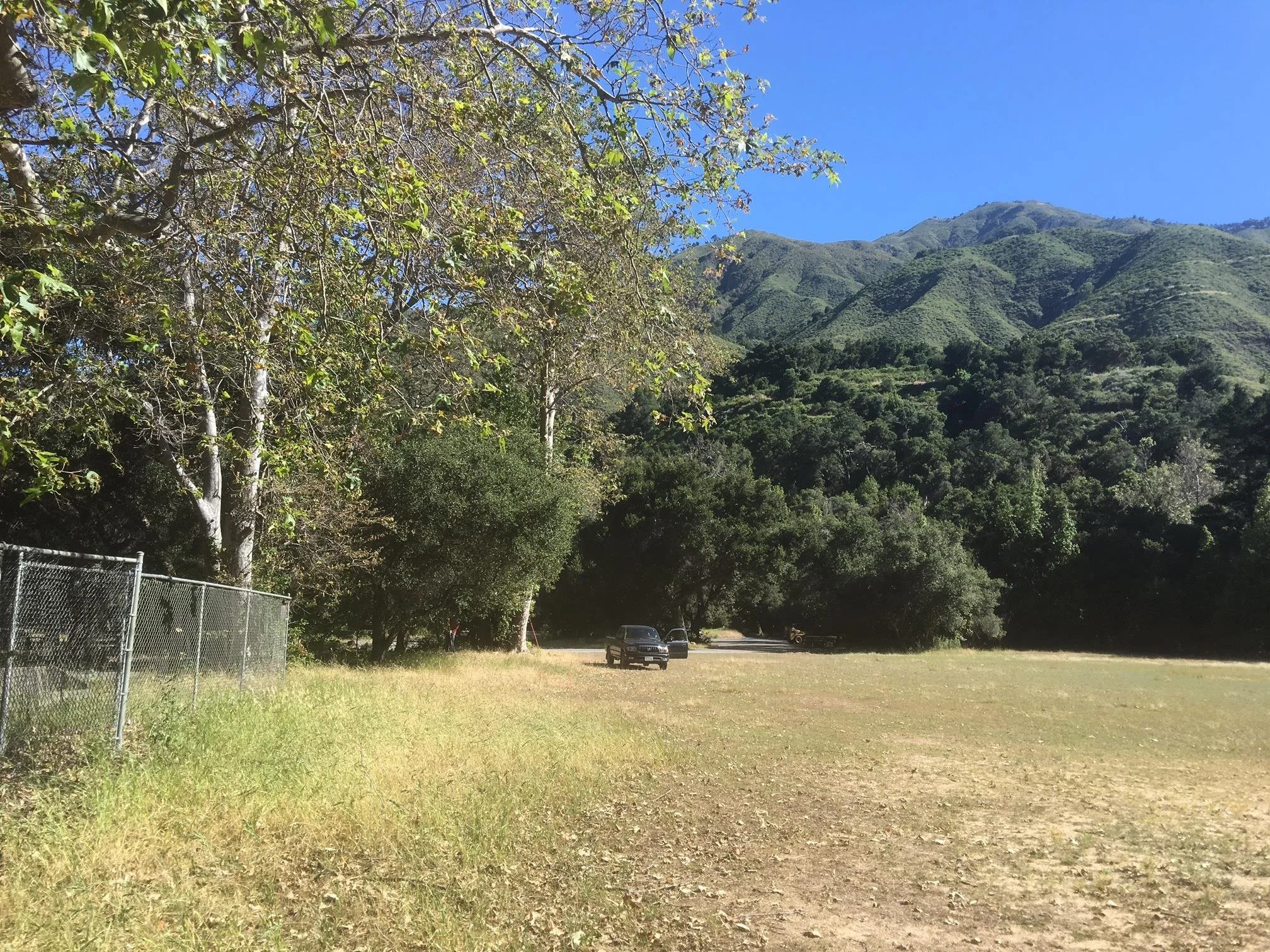 A grassy open area with a chain-link fence on the left, trees in the middle ground, and a mountain range in the background under a clear blue sky.