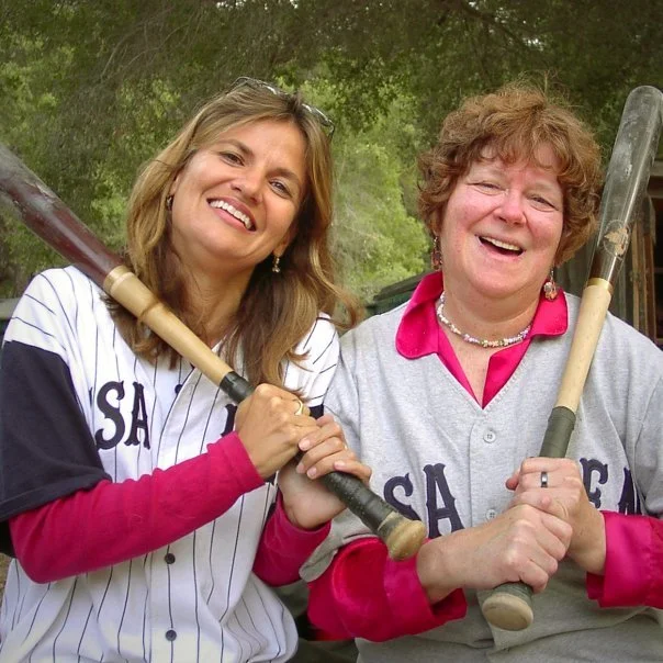 Two smiling women in baseball jerseys holding bats outdoors.