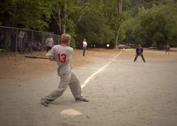 A baseball player wearing a gray uniform with the number 13 on the back is preparing to hit a baseball on a dirt field. Spectators are behind a chain-link fence, and other players are positioned at various points in the outfield. Trees line the backg