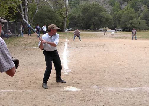 A person in a white shirt and hat swinging a baseball bat at a baseball in a park with other people in the background.