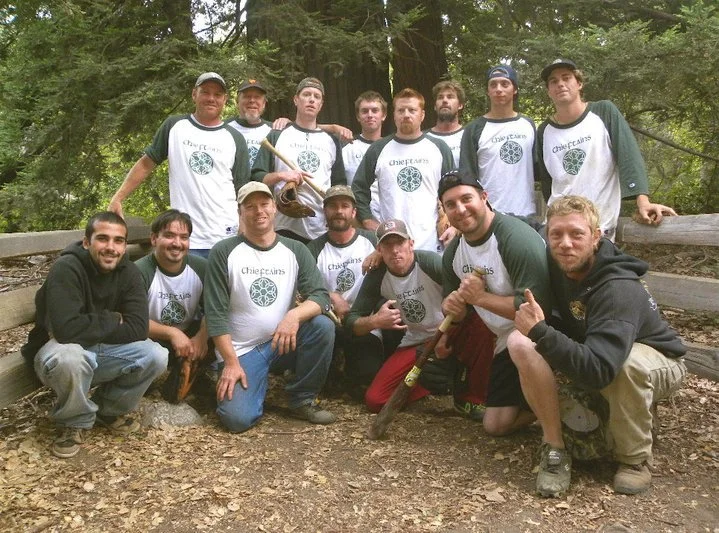 Group of men outdoors in a forest, some wearing matching shirts with a circular logo that says 'Chiefs.' They are smiling, posing for a photo, and some are holding tools, with a fallen log in the background.
