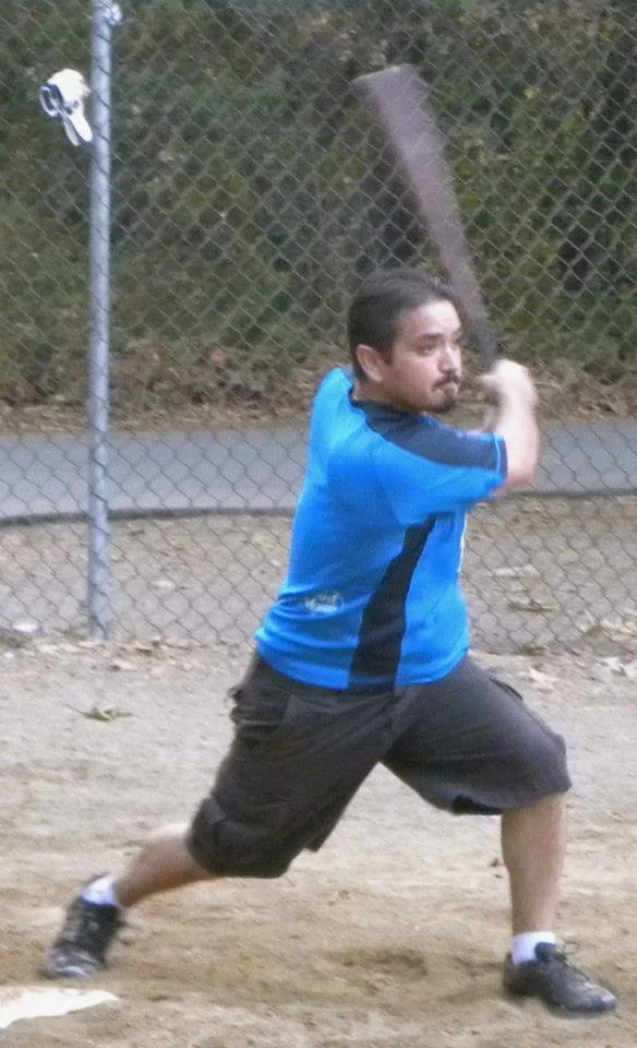 A man in a blue and black sports jersey and black shorts swinging a baseball bat on a dirt field, with a chain-link fence and trees in the background.