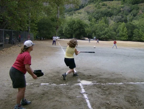 Children playing baseball on a dirt field surrounded by trees and hills.