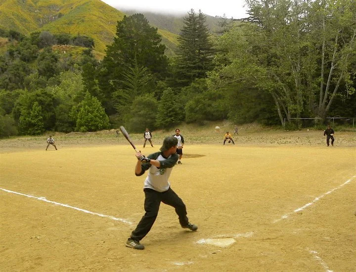 A person batting at a baseball game on a dirt field surrounded by trees and mountains in the background.