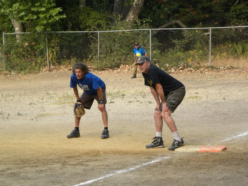 Two men in athletic gear are crouching on a baseball or softball field near a first base with one man wearing a blue shirt and the other in a black shirt. A person in the background is walking or stretching near the outfield fence with trees beyond.