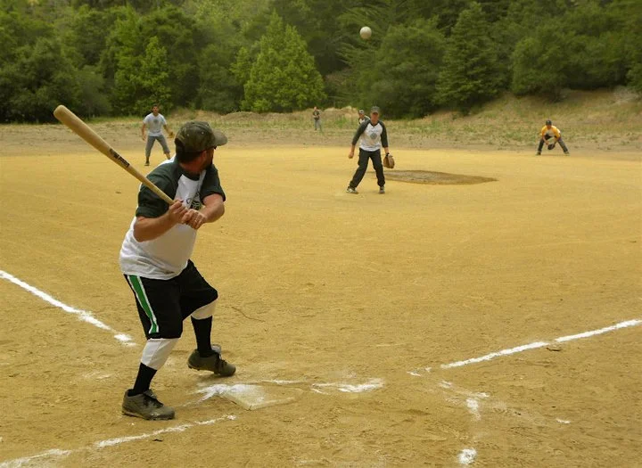 People playing baseball on a field surrounded by trees, with one player preparing to hit the ball.