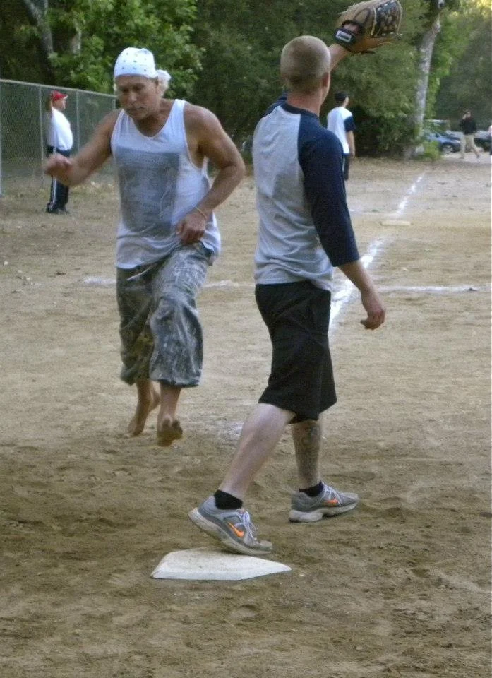 Two individuals playing a game on a dirt baseball or softball field. One person is middle-aged with gray hair, wearing a white sleeveless shirt, patterned shorts, and a white cap. The other is a young boy with short hair, wearing a gray and black shi
