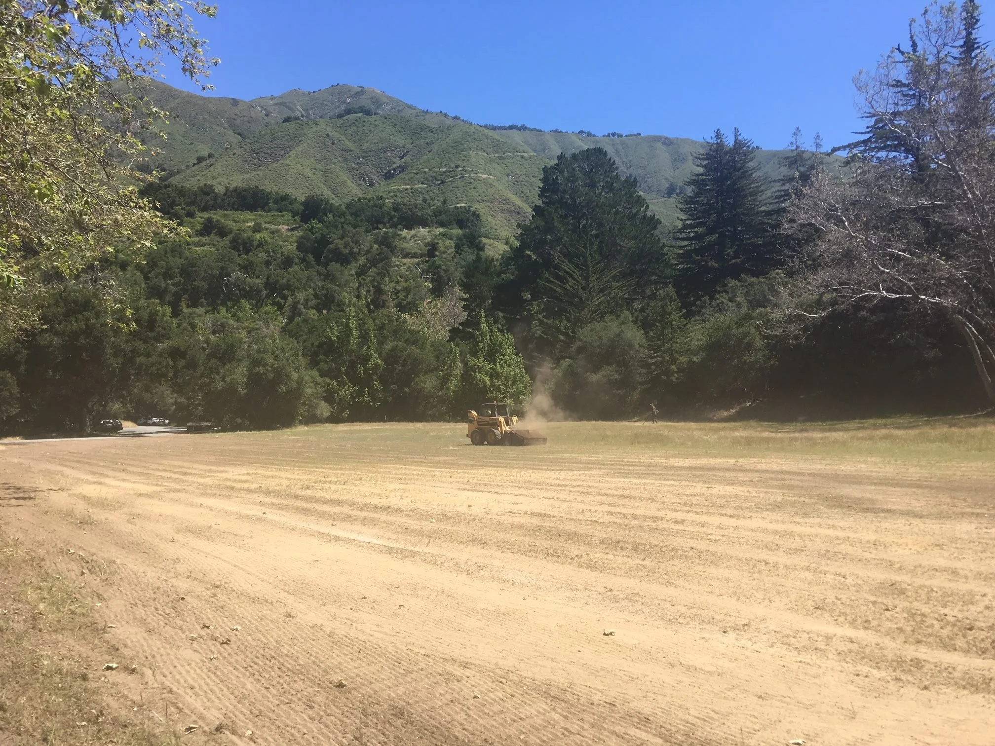A dirt or gravel parking lot with some cars parked on the left side and a bulldozer working on the ground. Tall trees surround the area, with lush green mountains in the background under a clear blue sky.