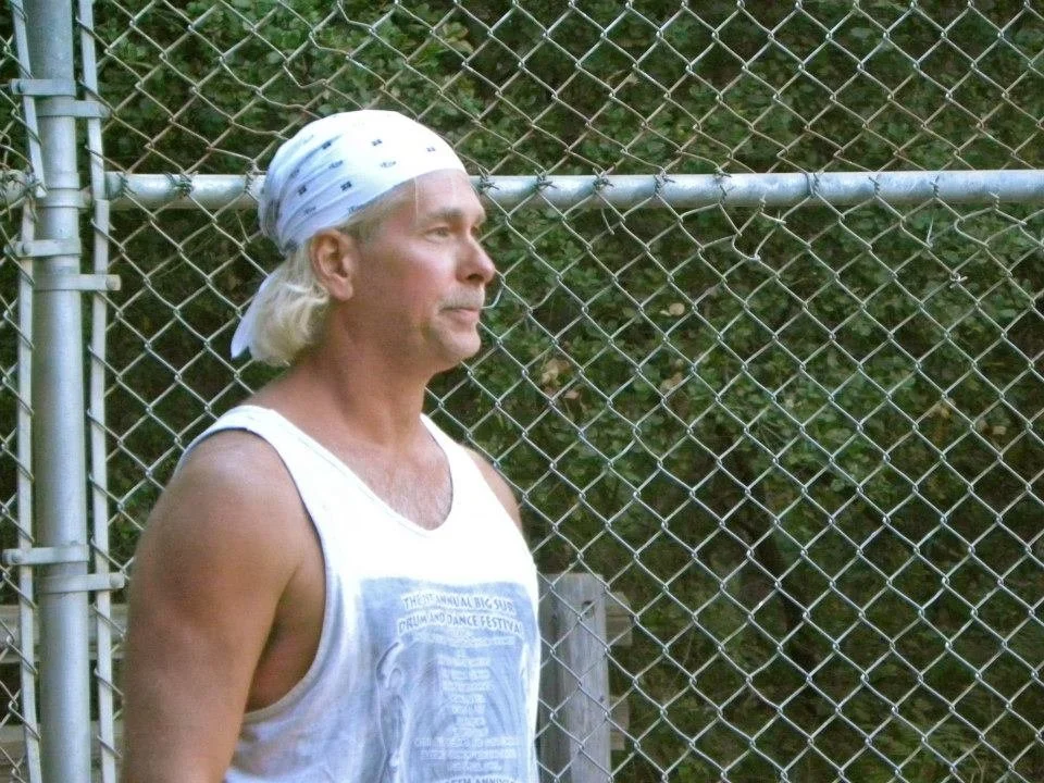 A middle-aged man with long blonde hair, wearing a white bandana and a sleeveless white shirt, standing in front of a chain-link fence with greenery behind it.