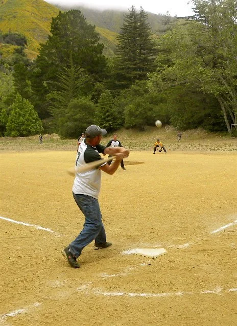 Person batting in a baseball game on a dirt field with trees and mountains in the background.