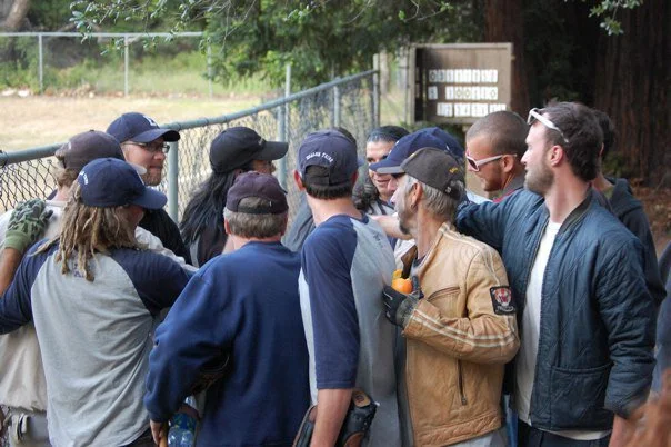 Group of people outdoors gathered near a chain-link fence, some wearing caps and sunglasses, engaging in conversation.