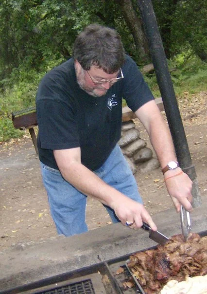 A man grilling meat outdoors at a park or backyard, using tongs to handle the food on a barbecue grill.