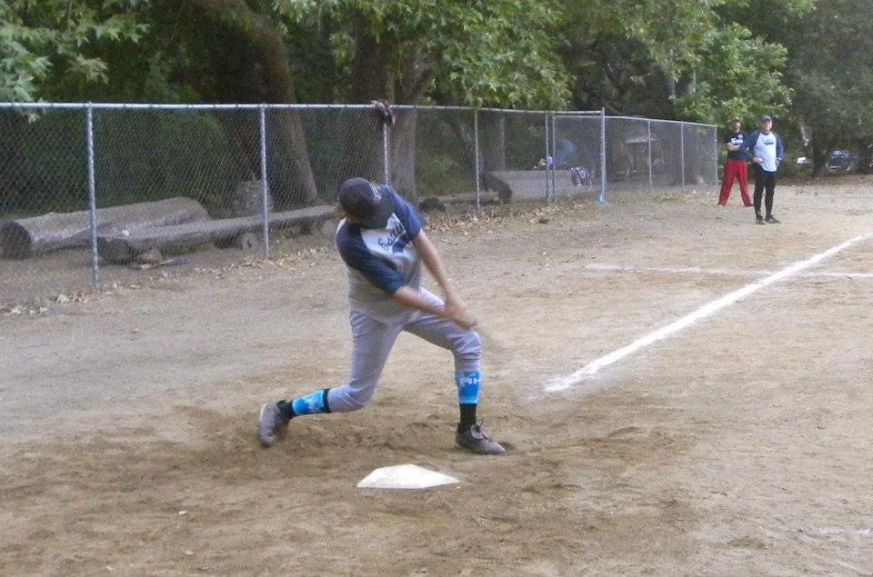 Child practicing baseball swing at a dirt field, with a chain-link fence and trees in the background, and adults observing in the distance.