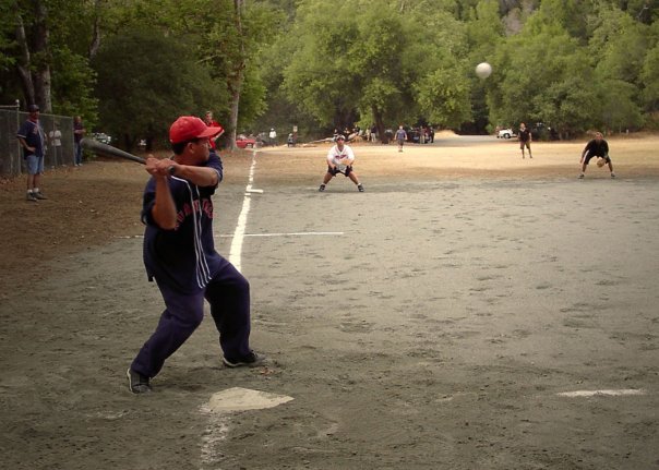 Young boy in a red cap and navy shirt batting in a softball game at a park.