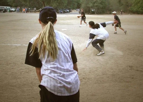 Kids playing baseball on a dirt field, with one girl in the foreground watching and others in action, surrounded by trees and parked cars.
