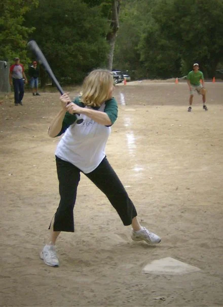 A woman with blonde hair batting at a baseball game in a park.