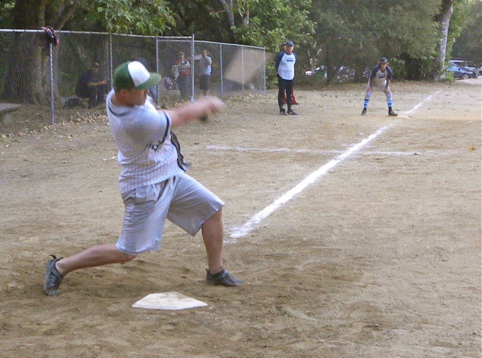 A young boy in a baseball uniform is swinging a bat at a pitched ball on a dirt baseball field, with other kids and spectators in the background.