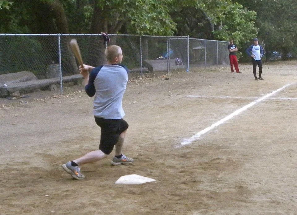 A young boy in a baseball uniform prepares to hit a baseball on a dirt field, with two other children watching from the outfield fence and trees in the background.
