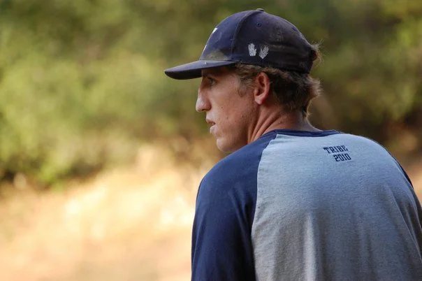 A young man wearing a dark cap and a blue and gray T-shirt looking off into the distance outdoors.