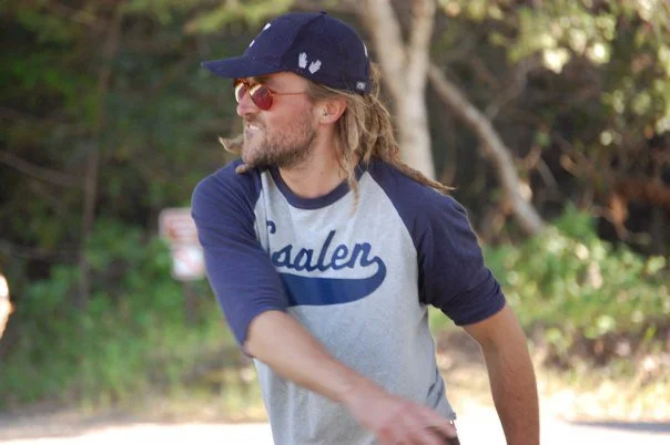 Man with long hair, beard, and glasses wearing a baseball cap and a gray and navy blue baseball tee, outdoors in a wooded area.