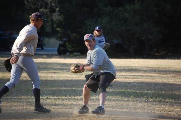 Three men playing baseball on a field, one with a glove ready to catch the ball and two others in casual sportswear.