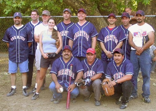 Group of baseball players in team uniforms and one woman in casual clothes, standing outdoors on a baseball field. Several players are wearing caps and holding baseball equipment.