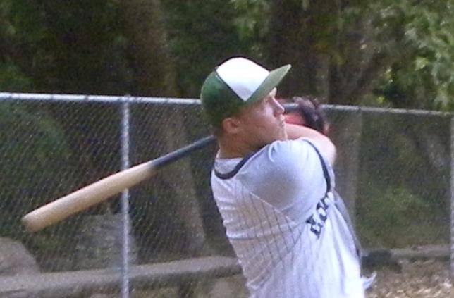 Person in a white baseball jersey and cap swinging a baseball bat in a park.