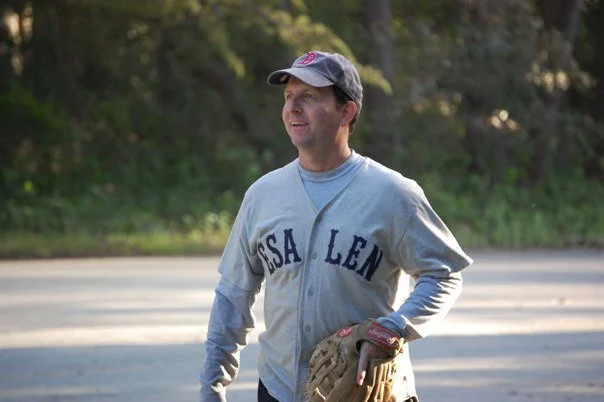 A man in a gray baseball jersey with 'USA LEN' written on it, wearing a baseball cap, gloves, and holding a baseball glove, standing outdoors on a sunny day.