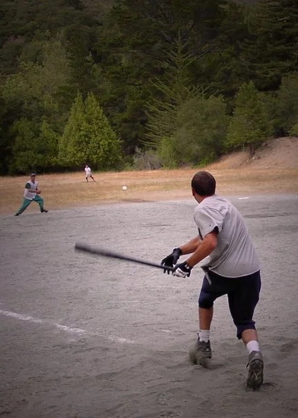 A person in gray shirt and black shorts playing baseball, preparing to swing at a pitch on a dusty baseball field surrounded by trees.