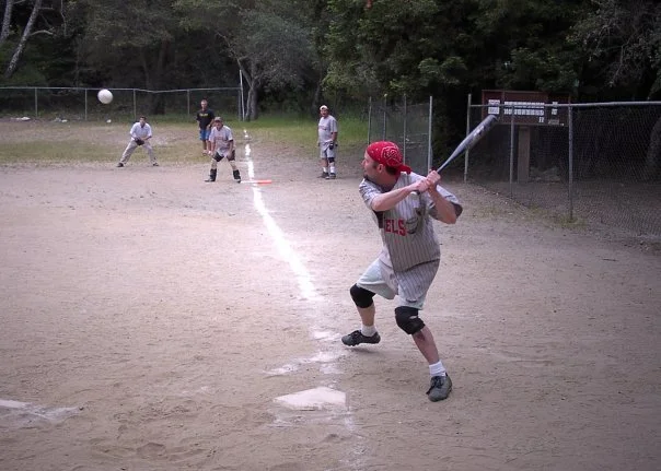 Young boy wearing a red helmet, gray pinstripe baseball uniform, and black knee pads swinging a bat at a baseball during a game on a dirt field.