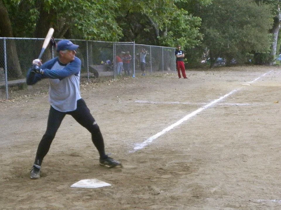 A person preparing to hit a baseball during a game on a dirt field, with other players and a fence in the background.