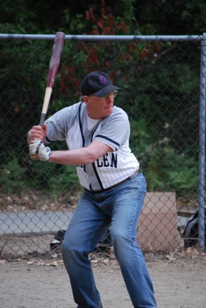 A man in a baseball uniform preparing to bat on a baseball field, with a chain-link fence and trees in the background.