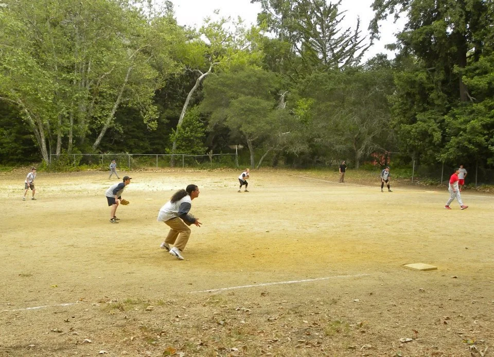 People playing baseball on a dirt field surrounded by trees.